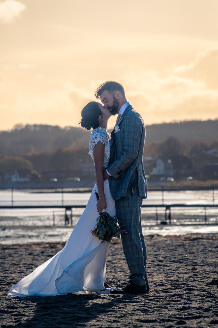 Couple kissing on the beach in the sun, embracing a romantic moment on their wedding day.