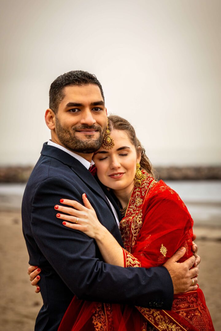 Bride hugging the groom at the beach with closed eyes, resting her head on his chest while he looks at the camera.