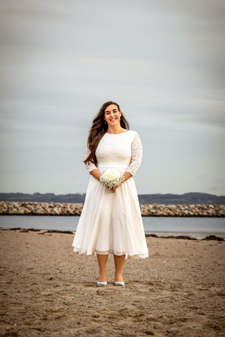 Full-body portrait of the bride smiling on the beach, holding a beautiful flower bouquet.