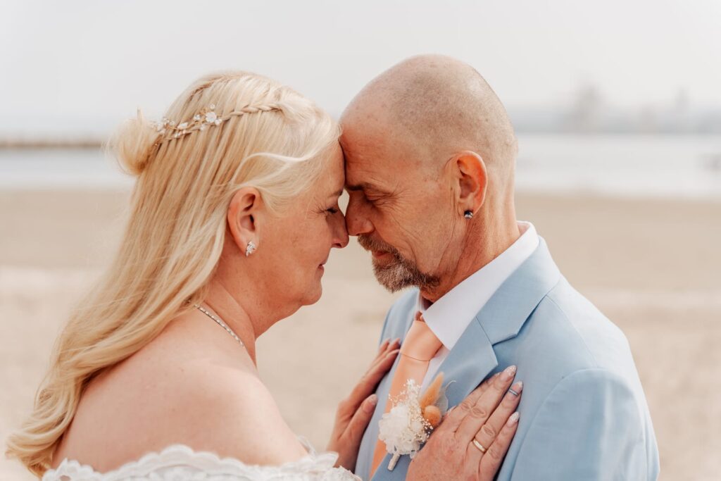 Couple leaning their foreheads together on the beach, her hand on his chest, in soft coastal light.