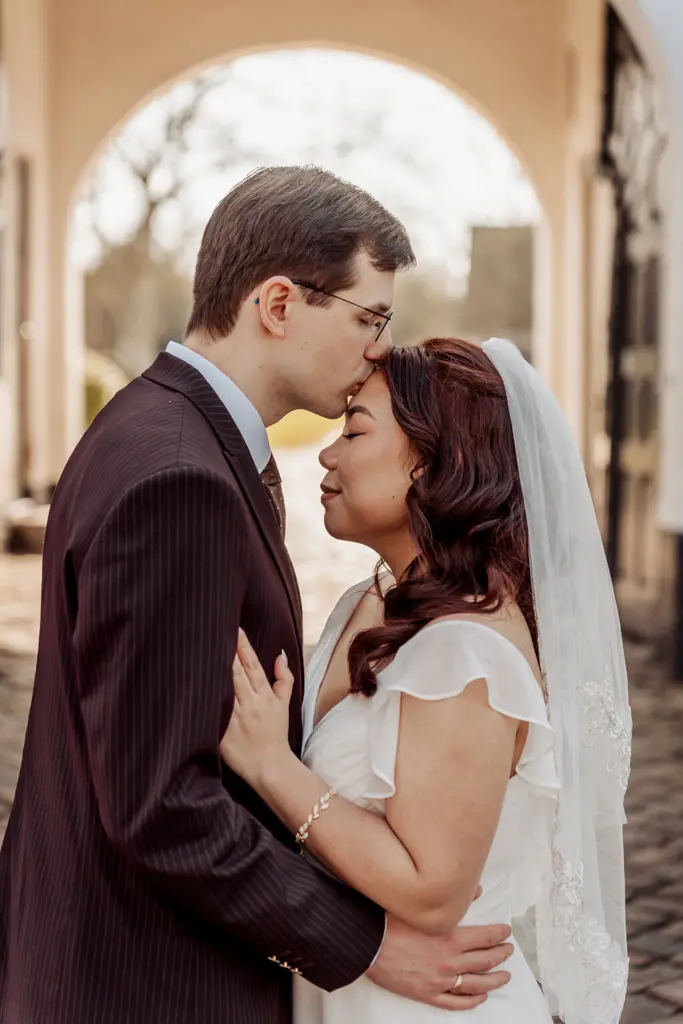 groom kissing the bride's forehead under a stone archway on a cobbled street