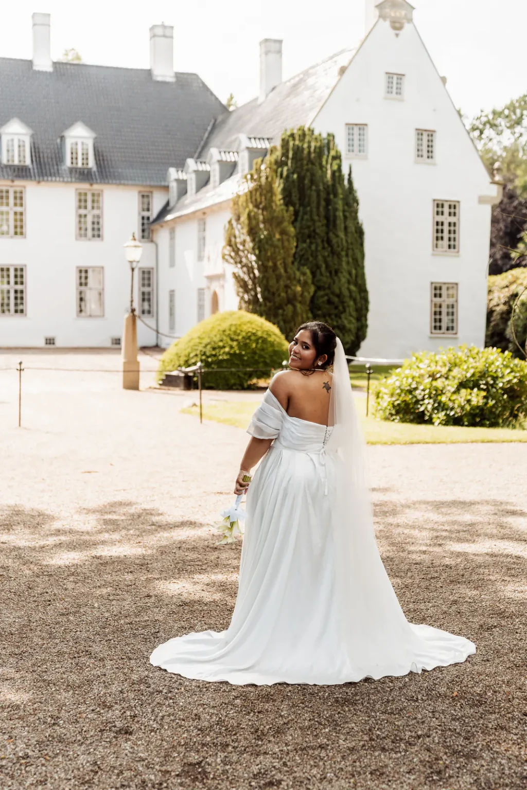 Bride smiling over her shoulder, her train spread across the gravel at Schackenborg Castle