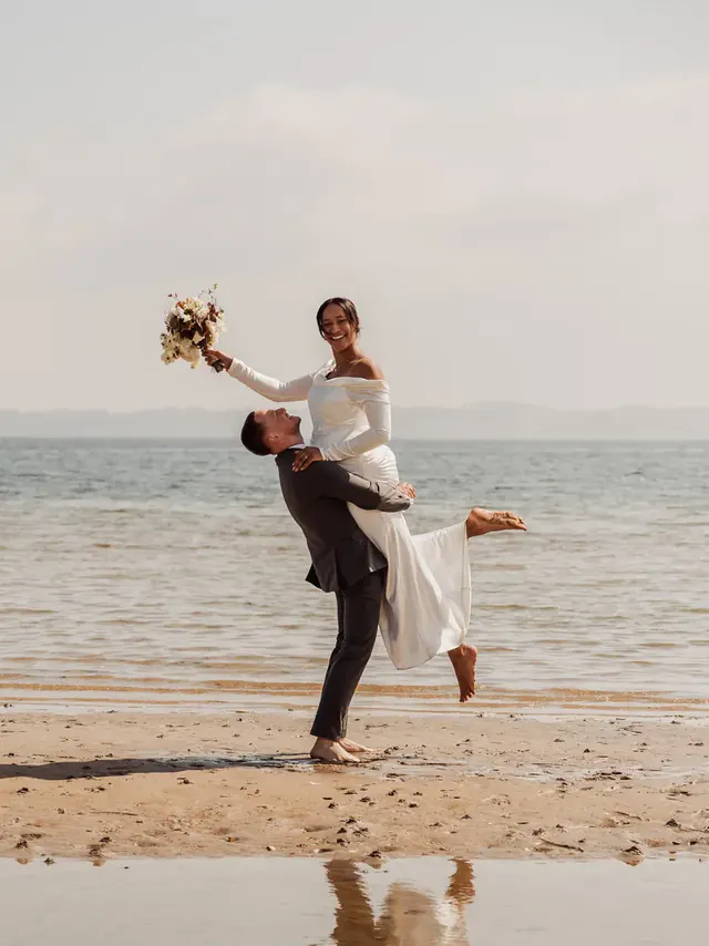 Groom lifting his bride on the beach in Aabenraa, her bouquet held high