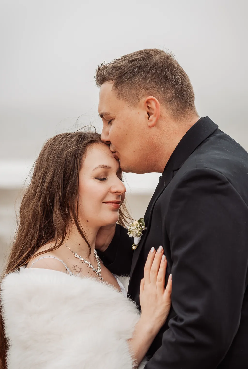 Groom kissing the bride on the forehead by the water on a misty winter day